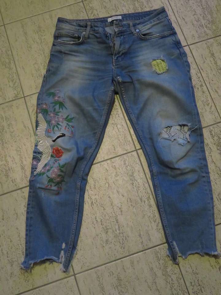 Pair of jeans with holes in the legs, distressed ankles, and a large embroidered area on the right leg
