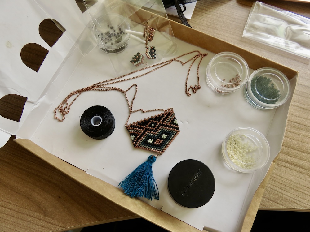 Small carboard box holding clear bead containers, a geometric beaded pendant, a spool of black thread and a clear plastic baggie containing smaller beaded pieces and copper-coloured studs